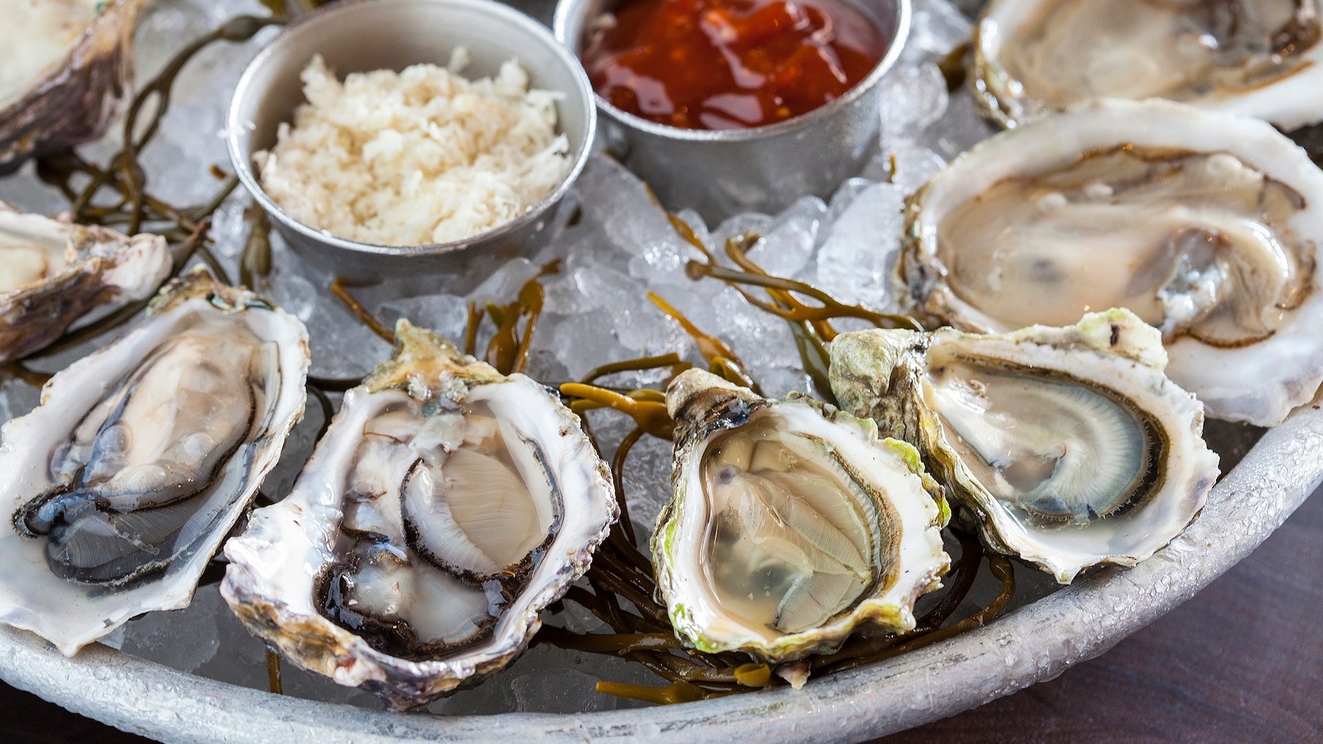 Close-up of a seafood platter with fresh oysters served on a bed of ice, accompanied by cocktail sauce, horseradish, and seaweed garnish.